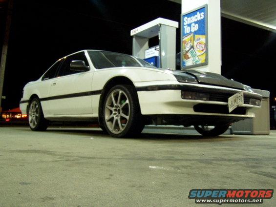 White coupe with aftermarket wheels and a black hood at a gas station