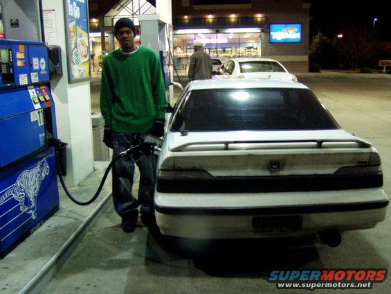 Guy in green sweater fueling a white sedan at a gas station at night. Another car is visible behind it