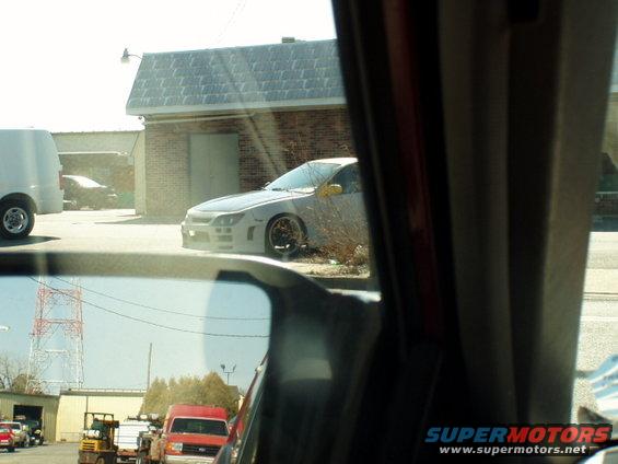 Gray car with aftermarket body kit parked near a brick building, seen from inside another vehicle