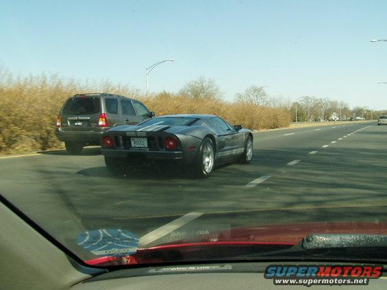 Dark grey Ford GT with white racing stripes on rear, driving on highway next to SUV. License plate reads 'RICES'