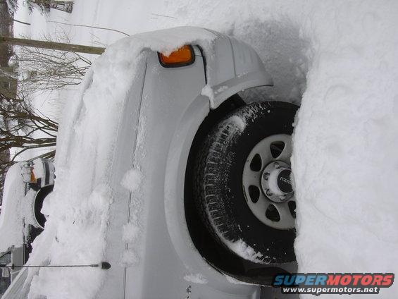 White truck buried in snow, only front corner and wheel visible. Another vehicle is partially visible to the left