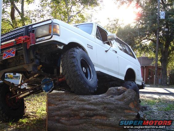 White 1989 Ford Bronco with off-road modifications driving over a large log