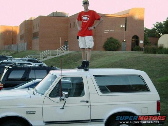 White SUV with a man standing on its roof, side view. The vehicle has a "BRONCO" badge