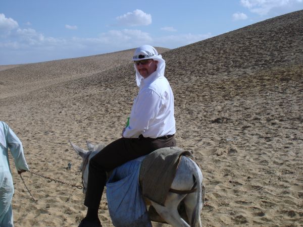 Man in white shirt and head covering rides a donkey in a sandy desert landscape with dunes under a blue sky