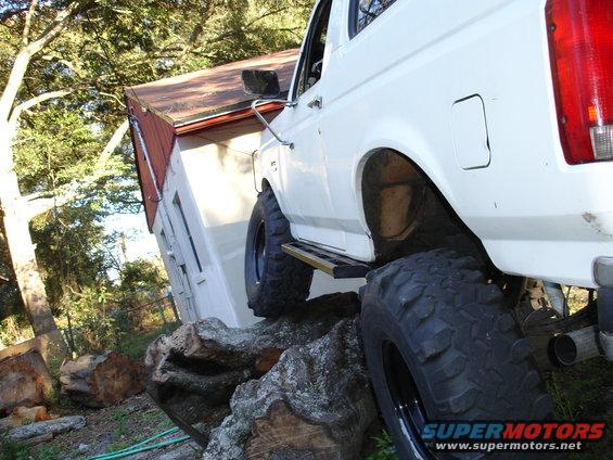 White Ford Bronco with large off-road tires on rocks near a small building