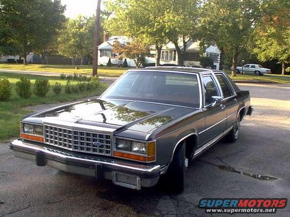 Dark gray 1987 Ford Crown Victoria LTD sedan parked on asphalt, front three-quarter view. Chrome grille and bumper. Another white car is visible in the background