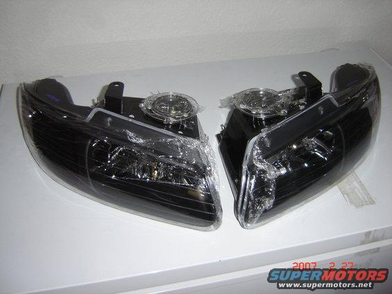 Two black aftermarket headlights, still in plastic wrap, on a white surface