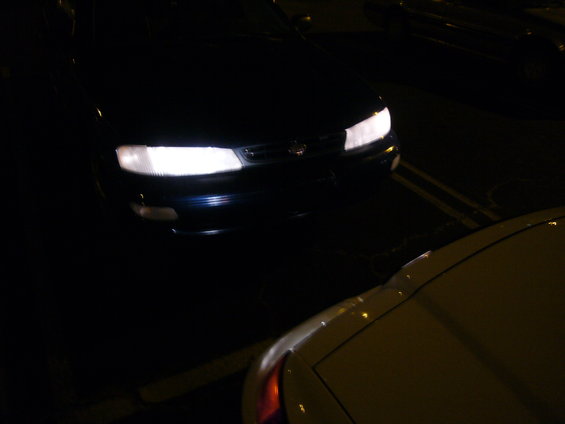 Headlights of a dark blue sedan at night, with another car in the foreground
