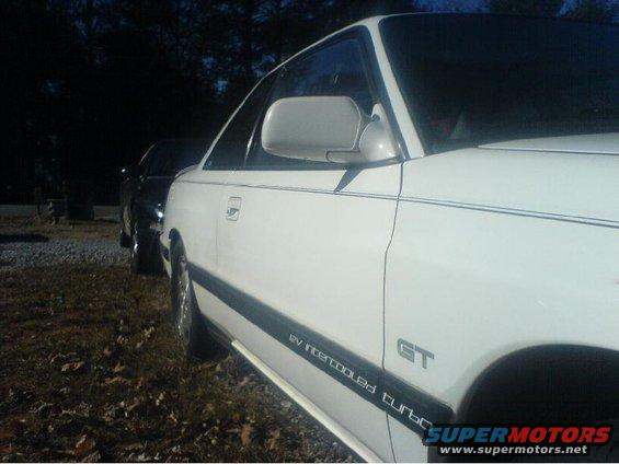 Side view of a white 1996 Ford Probe GT with '12v Intercooled Turbo' decal on the lower door trim. Another dark car is visible behind it