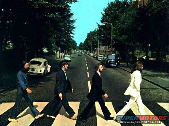 Four men walk across a zebra crossing on a street lined with trees and buildings, with several cars visible
