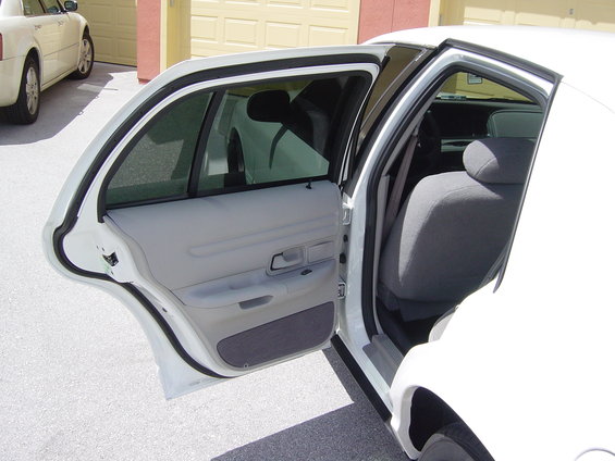 Open rear doors of a white sedan, showing gray cloth seats and interior panels. Another white car is parked nearby
