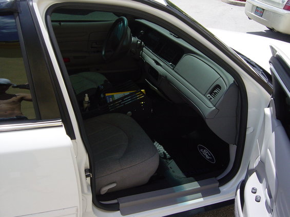 Interior of a white Ford Crown Victoria, showing the driver's seat, dashboard, and floor mats with a Ford logo