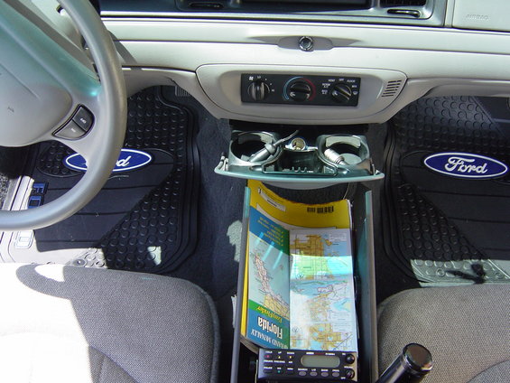 Car interior showing dashboard, steering wheel, and floor mats with Ford logos. A map and radio are in the center console