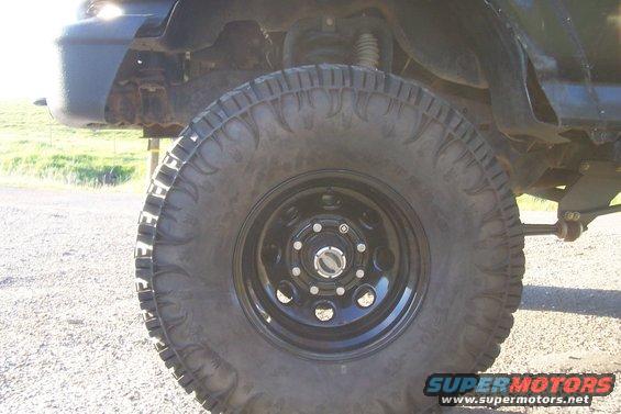 Black truck wheel with aggressive tread and black rim, showing suspension components and part of the fender