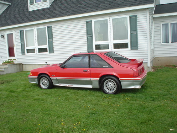 Red 1987 Ford Mustang GT coupe parked on grass in front of a white house