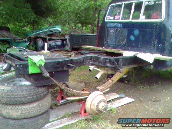 Truck chassis on jack stands, rear leaf springs visible, stacked tires, another green truck body in background