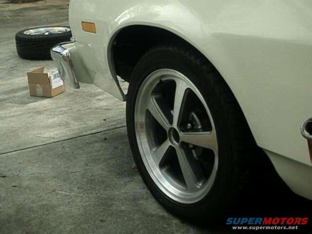 Close-up of a white car's side, showing a custom five-spoke wheel and tire. A spare tire and a small cardboard box are on the ground nearby