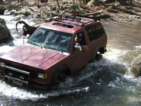 Red SUV with a roof rack driving through a rocky river, driver visible