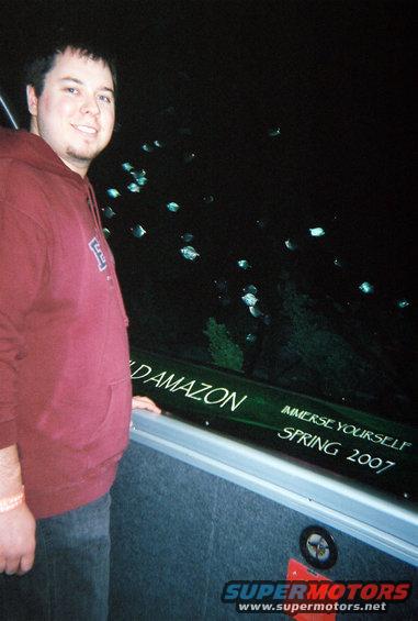 Man in maroon hoodie and jeans stands next to a dark green surface with text 'DAMAZON' and 'IMMERSE YOURSELF SPRING 2007'