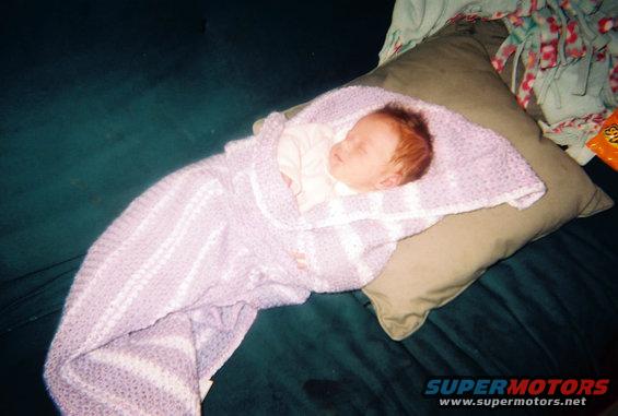 Baby sleeping on a pillow, wrapped in a purple and white blanket. A pillow and blankets are behind the baby