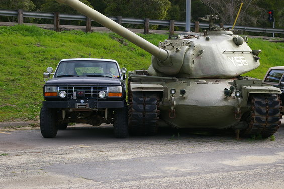 Ford truck parked next to a tank. The truck is a dark color with a white top and has a winch on the front
