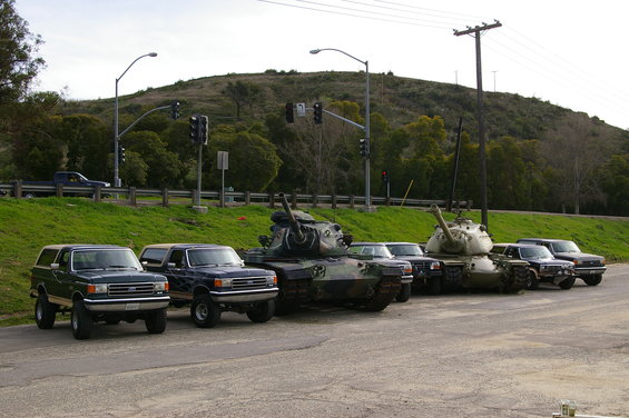 Two dark green Ford Broncos parked next to two military tanks, with more vehicles in the background