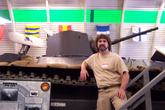 Man leans on a tank with sandbags in front. Display board reads 'Okinawa April 1945'