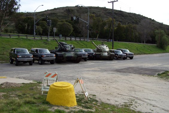 Two military tanks parked next to several SUVs on a dirt lot with a grassy hill in the background