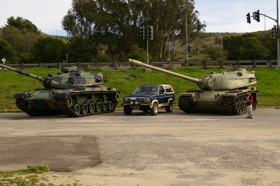 Blue SUV parked between two military tanks, front view