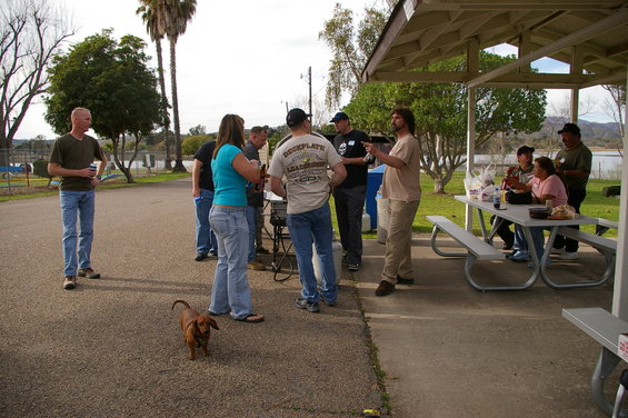 People gathered at picnic tables under a pavilion near a lake, with a dachshund in the foreground