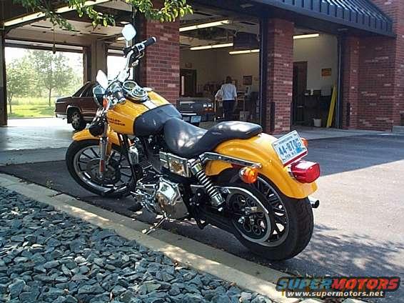 Yellow motorcycle parked on gravel, with a brown car in the background inside a garage