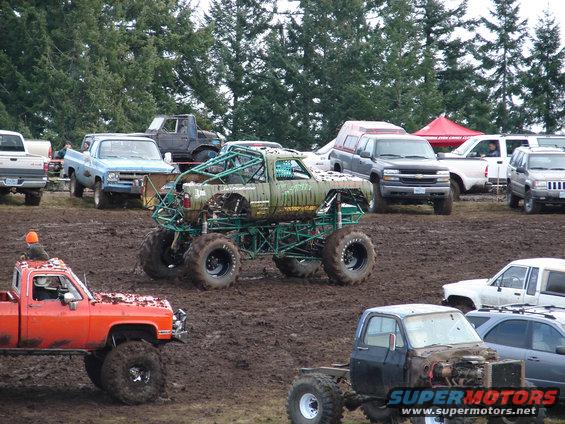 Mud bogging event with several trucks, including a green monster truck and an orange pickup. Other vehicles and trees in background