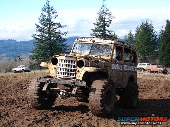 Mud-covered vintage Jeep wagon with large tires in a muddy field; white pickup truck and two SUVs in background