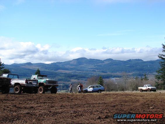 Muddy field with several trucks and a station wagon under a cloudy sky