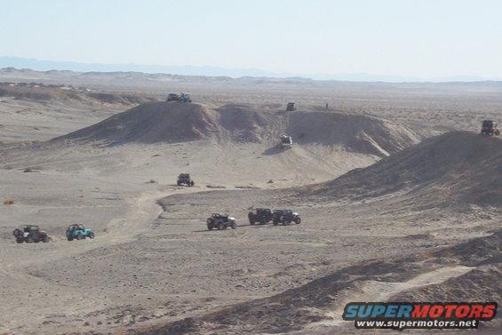 Several off-road vehicles are seen navigating sandy hills in a desert environment. One vehicle is on a crest, another descends a slope, and a group is gathered in the foreground