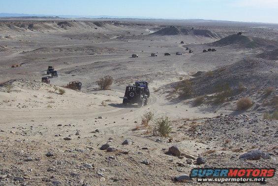 Several dark-colored off-road vehicles navigate a sandy desert trail under a clear blue sky