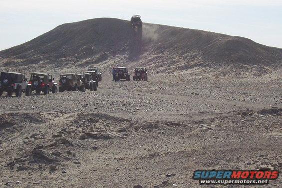Several Jeeps driving up a rocky hill in a desert landscape. One Jeep is near the top, kicking up dust