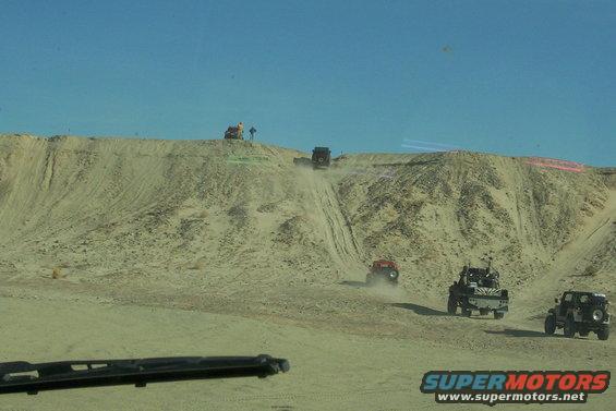 Several off-road vehicles driving up a sandy dune under a clear blue sky. One vehicle is kicking up dust as it climbs