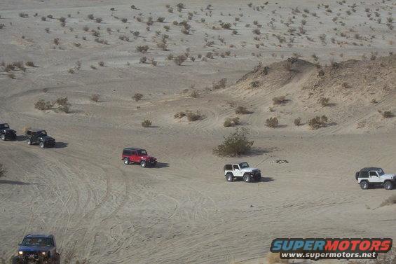 Several Jeeps driving on sandy terrain in a desert with sparse vegetation. Watermark reads SUPERMOTORS