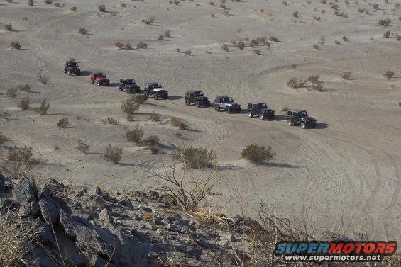 Several Jeeps driving in a line through a sandy, desert landscape with sparse vegetation