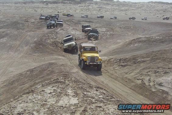 Yellow Jeep and white Jeep driving on sandy terrain with other Jeeps in the background