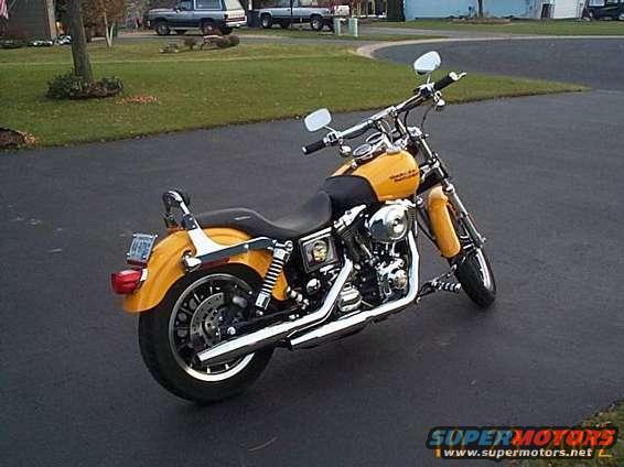 Yellow motorcycle parked on asphalt, rear three-quarter view. Chrome exhaust, black seat, license plate visible