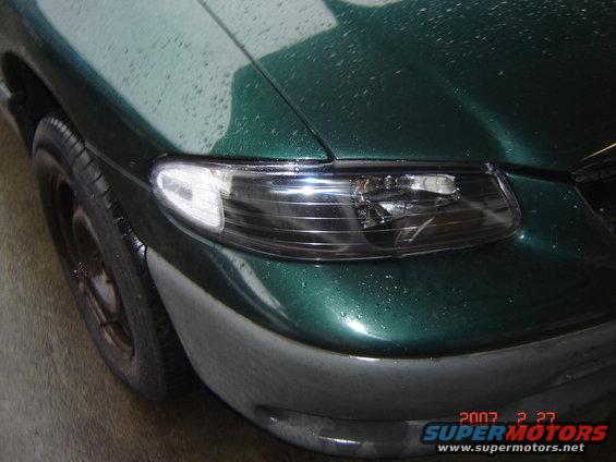 Front corner of a green minivan with water droplets on the paint. Shows the headlight and part of the tire