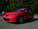 2007 Mazda 3 - A red 2007 Mazda 3 hatchback is parked on asphalt with a backdrop of trees. Another vehicle is visible to the left.