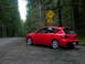 2007 Mazda 3 - A red 2007 Mazda 3 hatchback is parked on the side of a road in a forest, near a bike crossing sign.