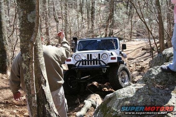 White Jeep Wrangler navigating a rocky, wooded trail. A person points ahead