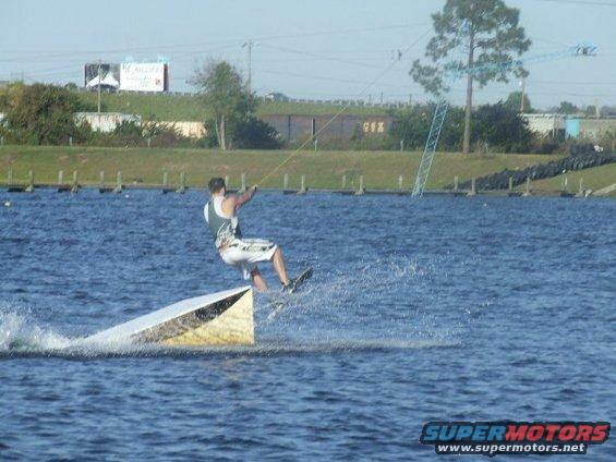 Wakeboarder performing a trick off a ramp in a lake, with a distant billboard and crane