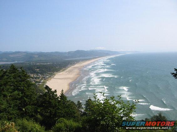 View from a hill overlooking a long sandy beach with waves crashing on the shore and mountains in the distance