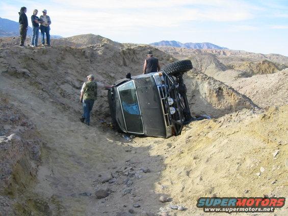 SUV flipped on its side in a sandy ravine with people standing around it