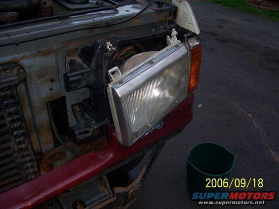 front end of vehicle with headlight removed, showing exposed wiring and rusty metal. Green bucket sits nearby